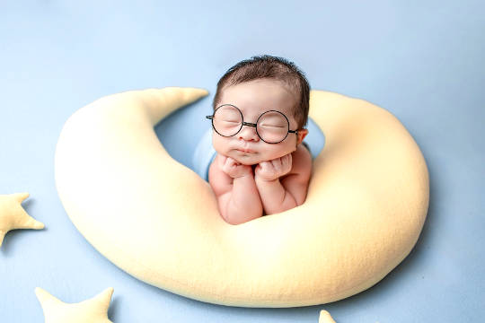 Re-member the Inner Child and Its Soul Mission baby with eyes closed wearing huge glasses and resting on a crescent moon-shaped pillos